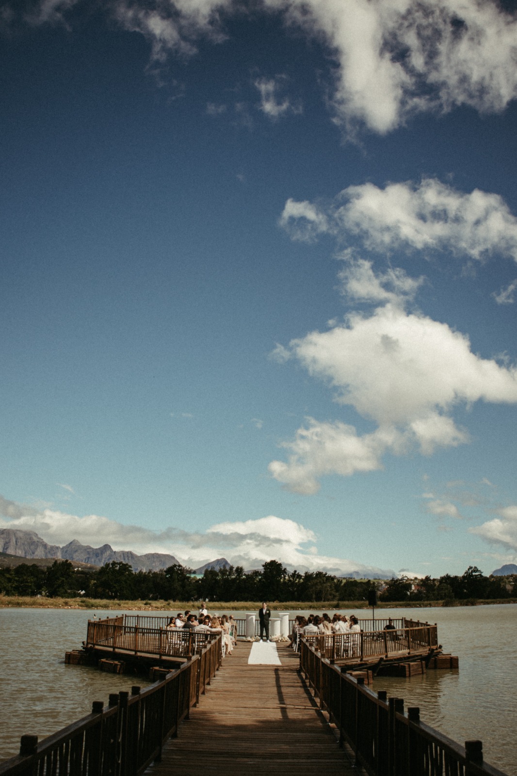 A wedding ceremony on a wooden deck over a lake with mountains in the distance